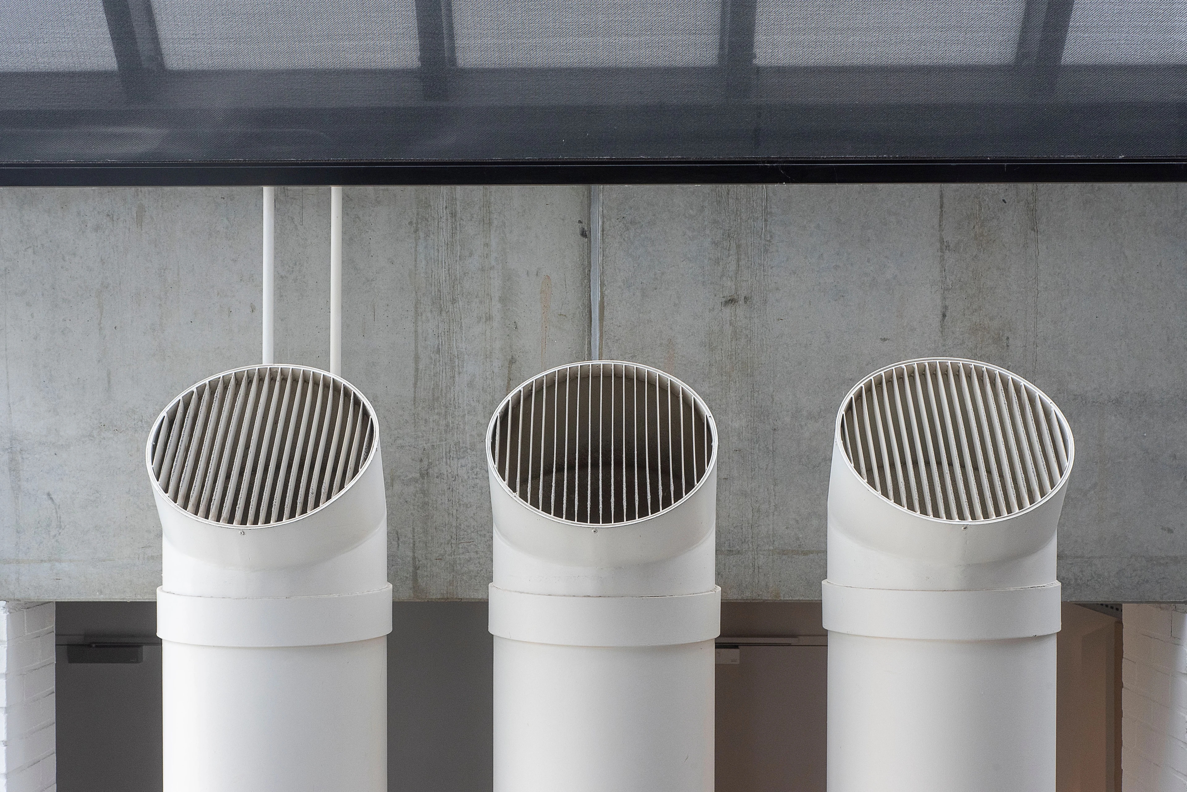 Três terminais brancos de ventilação predial com grelhas circulares, integrados a uma parede de concreto em ambiente arquitetônico; fotografia de referência.